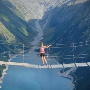 Eine Hängebrücke spannt sich über einen See, der von majestätischen Bergen umgeben ist. Die Brücke ist aus Holz und Seilen gefertigt, und ihre Struktur spiegelt sich im ruhigen Wasser des Sees. Im Hintergrund erheben sich die Berge mit ihren üppig bewaldeten Hängen und steilen Klippen. Der Himmel ist klar und blau, was die idyllische und friedliche Naturkulisse unterstreicht.